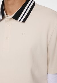 Pikétröja i ljusbeige med en svart och vit randig krage. Har en enhetlig tygtextur och en broderad logotyp på bröstet.