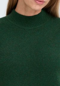 Pull en maille côtelée vert foncé avec un col haut. Le tissu texturé a une apparence douce, mettant en valeur un subtil mélange de teintes foncées.