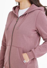 Pink zippered hoodie with a soft texture, adjustable drawstring hood, and front pockets. Features silver hardware and a small logo tag.