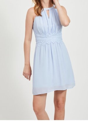 Femme portant une robe sans manches bleu clair avec un détail en dentelle à la taille et un petit décolleté en forme de goutte, debout devant un fond uni.