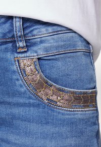 Denim jeans featuring a textured pocket with brown and gold sequins arranged in a decorative pattern, in a light blue wash with visible stitching.