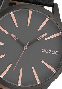 Cadran de montre noir mat avec un motif hexagonal, aiguilles et index en rose doré, et un bracelet en cuir noir. Marque "OOZOO."