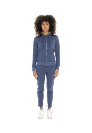 Une femme porte un sweat à capuche bleu zippé et un pantalon assorti, avec des baskets blanches, sur un fond blanc uni.