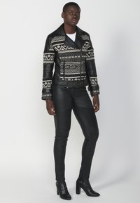 Giacca da motociclista in pelle nera con motivo a maglia geometrico, dotata di un ampio colletto e chiusura a zip, abbinata a pantaloni in pelle nera aderenti.