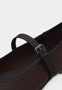 Chaussure Mary Jane marron foncé avec une bride réglable à boucle argentée et des coutures décoratives sur un matériau lisse.