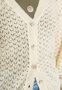 Kremowy kardigan dziany z ażurowym wzorem, dekoltem w serek i pięcioma okrągłymi guzikami, o fakturowanym wykończeniu i lekko luźnym kroju.