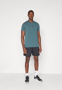 Camiseta deportiva de color verde azulado con mangas cortas y el logo de Adidas en negro, combinada con pantalones cortos negros y zapatillas deportivas negras, con acentos blancos.