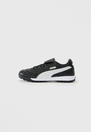 Black low-top soccer shoe with white side stripe, textured sole, lace-up front, and Velcro flap over laces on plain background.