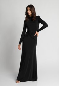 Kobieta w długiej, dopasowanej czarnej sukience z marszczeniami w talii, stojąca z jedną ręką na biodrze, a drugą luźno opuszczoną.