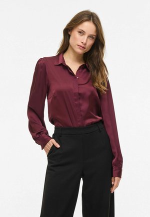 Kvinde med langt brunt hår iført en langærmet burgunderrød satinbluse, stoppet ned i højtaljede sorte bukser, poserer med den ene hånd i lommen.