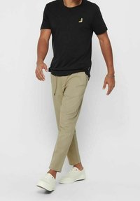 Schwarzes Kurzarm-T-Shirt mit einer kleinen gelben Bananengrafik, kombiniert mit beigefarbenen, schmal zulaufenden Hosen und weißen Sneakern.