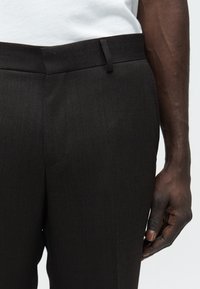 Το δεξί χέρι αναπαύεται δίπλα στη μέση, φορώντας σκούρο καφέ υφασμάτινο παντελόνι και ένα λευκό πουκάμισο μπαίνουμε μέσα.