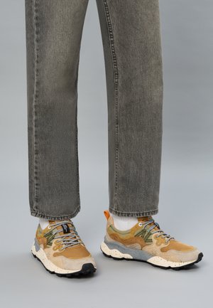 Person in grauen Jeans, weißen Socken und tanfarbenen, grünen und grauen Sneakers mit dicken gepunkteten Sohlen, die auf einem grauen Boden steht.