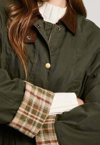 Veste olive avec un col en velours côtelé, fermeture éclair argentée et doublure de poignet à carreaux dans des teintes beige, verte et brune. Elle dispose d'une fermeture à bouton.