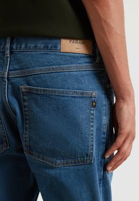 Jeans in denim blu con gamba dritta, caratterizzati da un leggero sbiadimento, cuciture a contrasto e un'etichetta in pelle beige con la scritta "FARAH" sulla vita.