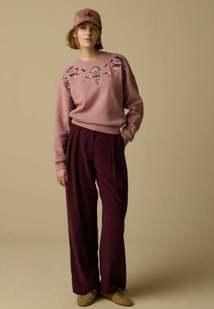 Femme portant un sweat-shirt rose à motifs floraux, un pantalon bordeaux à jambes larges, une casquette rose et des chaussures marron, debout avec une main dans la poche sur un fond beige.
