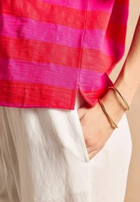 Bavlnený top s ružovými a červenými vodorovnými pruhmi. Má bočný rozparok a textúrovanú látku, v kombinácii s bielymi nohavicami a zlatými náramkami.