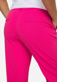 Pantalons ajustés roses avec une texture lisse, dotés de deux poches arrière et d'un design cintré. Le tissu semble léger et extensible.