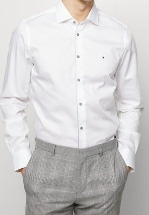 Uomo con camicia bianca a maniche lunghe abbottonata e pantaloni grigi a quadri, mani in tasca, in piedi davanti a uno sfondo chiaro e semplice.