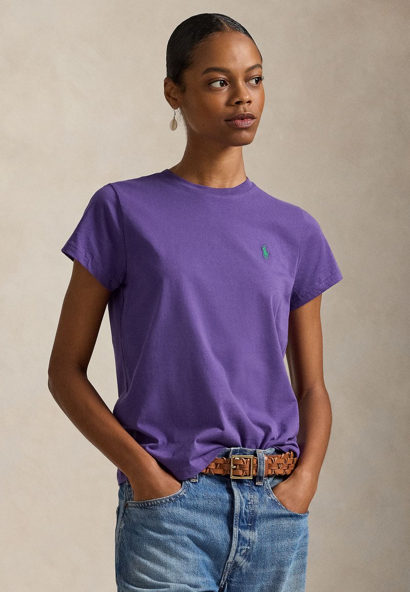 Μωβ βαμβακερό T-shirt με κοντά μανίκια, διαθέτει ένα μικρό πράσινο λογότυπο στο αριστερό στήθος, συνδυασμένο με μπλε τζιν και πλεκτή καφέ ζώνη.