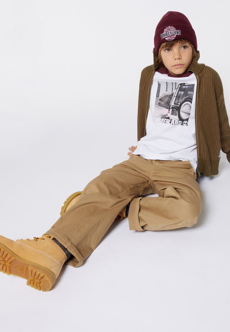 Cardigan marron côtelé, t-shirt graphique blanc, pantalons khaki et bottes jaunes avec semelles texturées. Bonnet en maille bordeaux avec détail de logo.