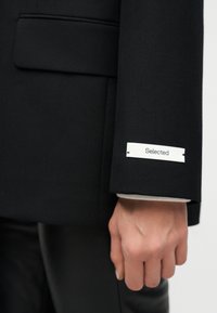 Χέρι που φορά μαύρο σακάκι με λευκή ετικέτα "Selected" κοντά στον καρπό στο μανίκι, μπροστά σε μονόχρωμο φόντο.