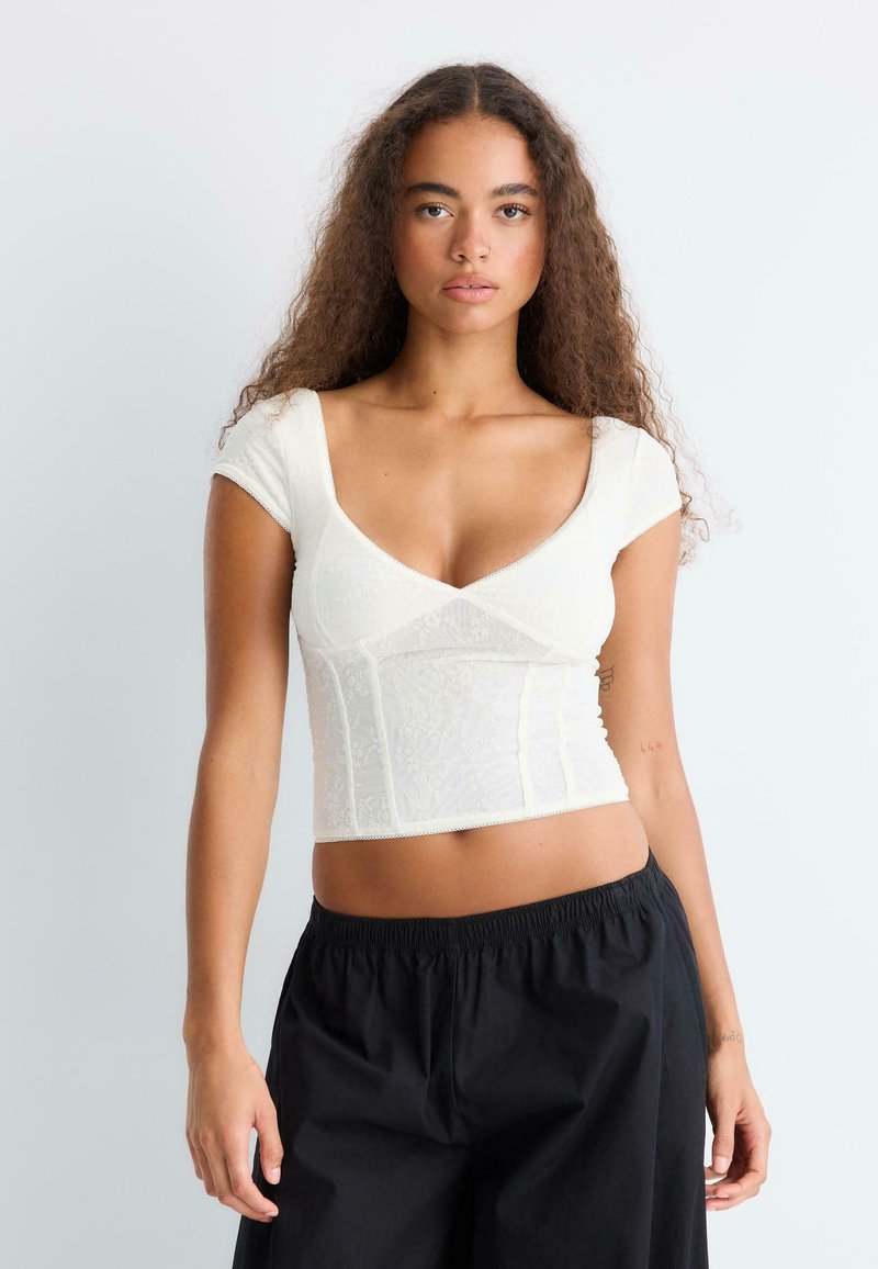 Femme aux longs cheveux bouclés porte un haut blanc ajusté à manches courtes et un pantalon noir ample, debout devant un fond gris clair.