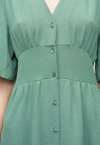 Πράσινο φόρεμα με υφή, κοντά μανίκια, εφαρμοστή μέση, V-λαιμόκοψη και μια κάθετη σειρά κουμπιών στο κέντρο μπροστά.