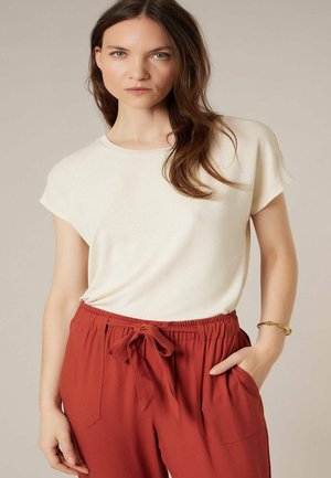 Femme portant un haut crème à manches courtes et un pantalon à cordon de couleur rouille, debout avec une main dans sa poche, sur un fond uni.