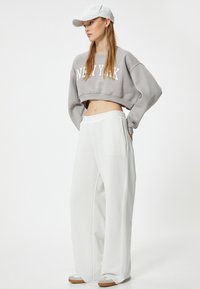Felpa grigia corta con la scritta "NEW YORK", abbinata a pantaloni bianchi larghi. Il modello indossa un cappellino bianco e calzature chiare.