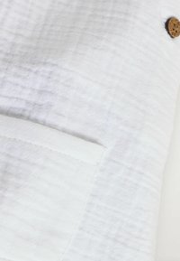 Weißer, strukturierter Stoff mit einer vorderen Tasche und einem runden Holzknopf-Detail. Das Design ist einfach und minimalistisch.
