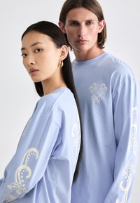 Light blue long-sleeve shirts feature floral designs in white on the sleeves and a central graphic. The fabric appears soft and casual.