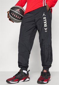 Black, lightweight, jogger-style pants with elastic cuffs, featuring white "BULLS" text and logo. Paired with red and black athletic shoes.