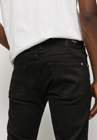 Persona vestida con jeans negros y una camiseta blanca, vista desde atrás, mostrando los bolsillos traseros y una etiqueta de logo en la cinturilla.