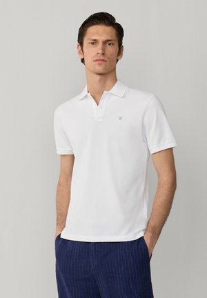 Ung mand iført en hvid kortærmet polotrøje og marineblå bukser med striber, stående med hænderne i lommerne foran en ensfarvet baggrund.