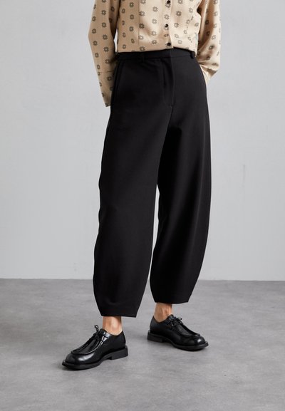 Pantalons noirs fuselés avec une texture lisse, dotés d'une taille mi-haute et de poches, associés à des chaussures en cuir noires à lacets.