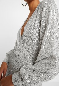 Silverglittrig klänning med djup V-ringning och långa, puffiga ärmar. Tyget har en texturerad, skimrande yta med en figurnära siluett.