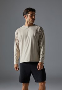 T-shirt de sport à manches longues de couleur beige clair avec un col rond et des panneaux texturés, associé à un short noir. Tissu doux et léger.