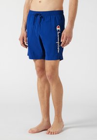 Shorts de bain bleus en tissu léger, dotés d'une taille élastique avec cordon de serrage, et d'un logo "Champion" blanc sur le côté.