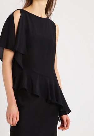 Femme portant une robe asymétrique noire avec une manche courte et un ourlet froncé sur un côté, sur un fond clair uni.