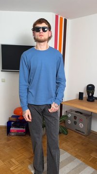 Sininen pitkähihainen paita, jossa on teksturoitu kangas, yhdistetty harmaisiin liehuviin farkkuihin. Asusteina mustat aurinkolasit ja näkyvät tatuoinnit käsissä.