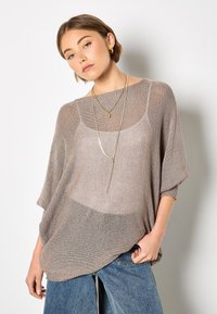 Leichter, taupefarbener Strickpullover mit lockerem Schnitt, weiten Ärmeln und einem strukturierten Muster, kombiniert mit blauen Jeanshosen und geschichteten Halsketten.