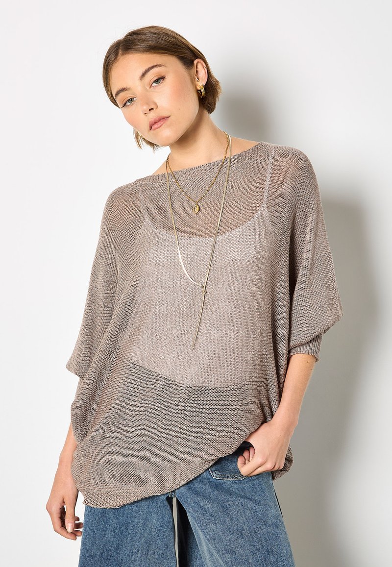 Leichter, taupefarbener Strickpullover mit lockerem Schnitt, weiten Ärmeln und einem strukturierten Muster, kombiniert mit blauen Jeanshosen und geschichteten Halsketten.