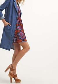 Un manteau en denim sur une robe fleurie colorée, présentant des motifs vibrants. Des talons compensés marron avec une base en liège, mettant en valeur un design unique.