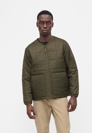 Homme portant une veste matelassée vert olive avec des poches avant sur une chemise noire et un pantalon beige, sur un fond blanc uni.