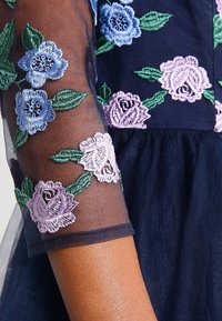 Marine jurk met doorschijnende, geborduurde mouwen met blauwe en lavendelkleurige bloemen en groene bladeren. Gelaagd stof en aansluitende lijfje.