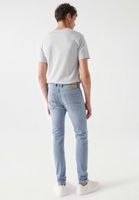 Vaqueros de color azul claro con un corte ajustado, bolsillos traseros y una etiqueta de logo marrón en la cintura, combinados con una camiseta gris y zapatillas blancas.