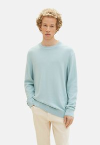 Pullover leggero di colore azzurro in maglia con scollatura rotonda e maniche lunghe, caratterizzato da polsini e orlo a coste, indossato sopra pantaloni color crema.