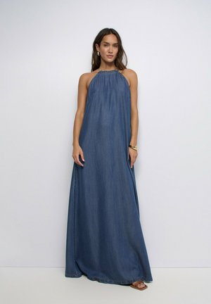 Mujer con vestido maxi de mezclilla azul sin mangas, de pie frente a un fondo claro y sencillo, complementado con pendientes, pulsera y sandalias.