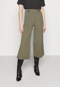 Kvinna i olivgröna vida cropped byxor med svarta knappar i midjan, kombinerat med svarta högklackade knähöga stövlar och svart topp.